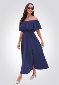 Abito maxi blu mare con scollo off-the-shoulder, volants al collo, vita elastica e uno spacco laterale. Il modello indossa sandali bianchi con i lacci.