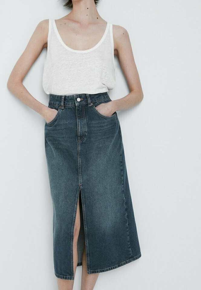 Femme portant un haut blanc sans manches et une jupe midi en denim foncé taille haute avec une fente sur le devant, mains dans les poches sur fond blanc uni.