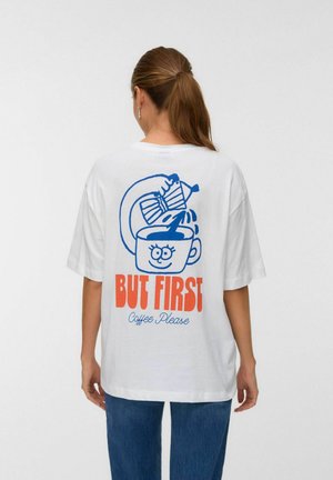 Witte katoenen t-shirt met een oversized fit. Grafisch ontwerp toont een blauwe koffiebeker en een gietkan met oranje tekst: "MAAR EERST Koffie, alsjeblieft."