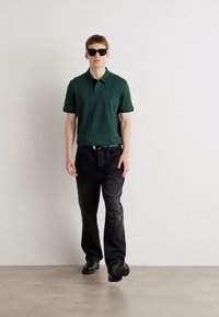 Zelený polo vrch, čierne džínsy a čierne topánky. Model má na sebe slnečné okuliare a stojí na neutrálnom podlaží pri bielej stene.