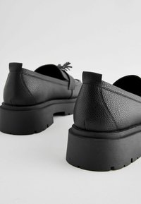 Black leather shoes with a textured finish, low heel, and rounded back. Features a minimalistic design and rubber sole with ridges.