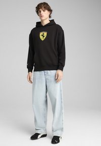 Sort hættebluse med Ferrari-logo, frontlomme og ribbede mansjetter, parret med lyseblå vide jeans og sorte sneakers.