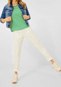 Woman wearing a green and white striped shirt, denim jacket, cream pants, and beige sneakers standing with one leg lifted against a plain background.
