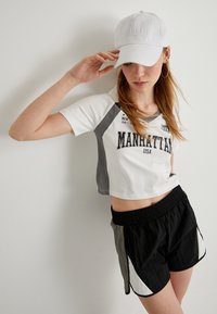 Beskaaret hvid og grå T-shirt med "MANHATTAN USA" print, parret med sorte shorts med grå og hvide paneler og elastisk talje. Hvid kasket.