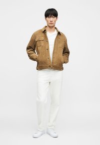 Άνδρας που φοράει καφέ σουέτ μπουφάν πάνω από λευκό πουκάμισο, λευκό παντελόνι και λευκά αθλητικά παπούτσια, στέκεται με τα χέρια στις τσέπες του μπουφάν.