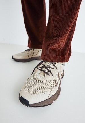Personne portant des baskets beiges et marron avec des lacets foncés et un pantalon en velours côtelé marron, debout sur une surface claire.