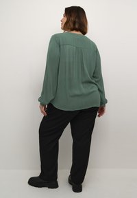Blouse verte légèrement texturée avec des manches longues, associée à un pantalon noir ample et des chaussures noires épaisses. Vue de dos, design simple, sans motifs.