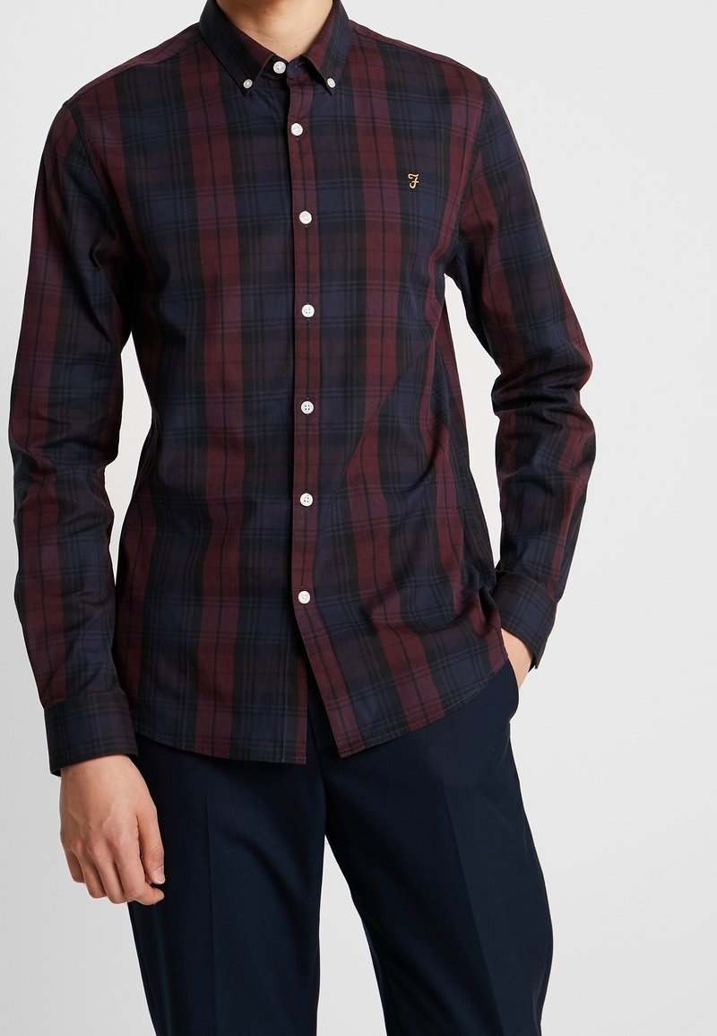 Chemise à carreaux originale en rouge foncé et bleu marine. Dispose d'un col boutonné, de manches longues et d'un petit logo brodé sur la poitrine.