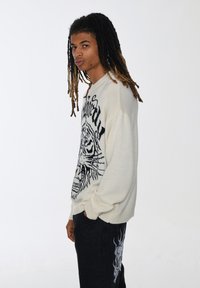 Maglione bianco a maglia con grafica di tigre nera. Vestibilità comoda, polsini a coste, indossato con jeans scuri caratterizzati da un design di teschio a motivi.