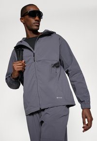 Giacca a vento grigia con zip frontale, tasche laterali, polsini elasticizzati e cappuccio. Indossata con una maglietta nera e una backpack.