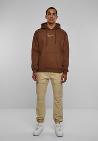 Felpa marrone con cappuccio, fascette e tasca frontale, abbinata a pantaloni cargo beige con tasche laterali e scarpe da ginnastica bianche.