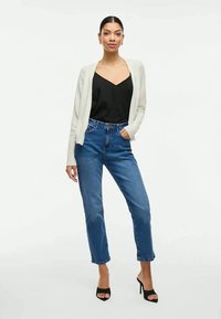 Cardigã cinza claro sobre um top preto, combinado com jeans de cintura alta azuis e sapatos de salto abertos pretos. Texturas suaves, ajuste casual.