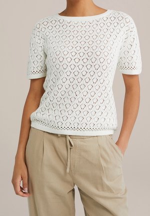 Vrouw draagt een wit gebreid topje met korte mouwen en een diamantvormig oogjespatroon, gecombineerd met beige broek met trekkoord, poseert met één hand in de zak.