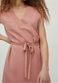 Robe rose poudré en tissu texturé, présentant un décolleté en V, des manches courtes et une ceinture nouée à la taille pour une silhouette définie.
