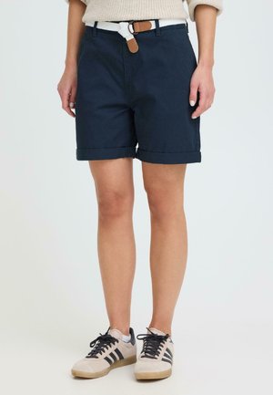 Persona con pantalones cortos azul marino con dobladillo enrollado, cinturón blanco tejido, suéter beige y zapatillas beige con franjas negras.