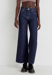 Jeans a gamba larga in denim scuro con vita alta, tasche frontali e cuciture a contrasto, abbinati a stivaletti neri alla caviglia.