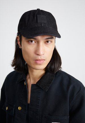 Gorra de mezclilla negra con parche de logo en la parte delantera, visera plana y correa ajustable. Se lleva con una chaqueta de pana negra con cuello y botones dorados.