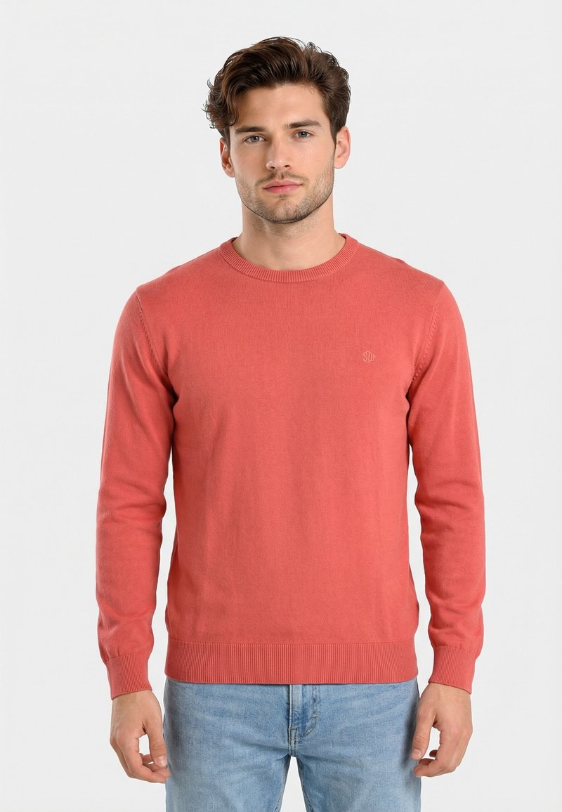 Uomo che indossa un maglione corallo a maniche lunghe semplice e jeans azzurri, in piedi davanti a uno sfondo bianco semplice.