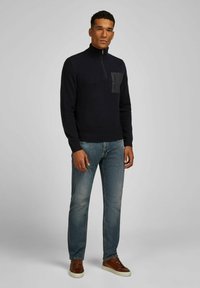 Marineblauwe pullover trui met hoge kraag, ritssluiting en een zwarte borstzak. Gecombineerd met lichtblauwe jeans en bruine sneakers.