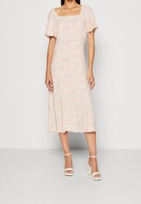 Mujer con un vestido midi floral en tonos pastel, con mangas cortas tipo volantín y escote cuadrado, combinado con sandalias de cuña blancas.