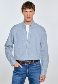 Camisa de cuadros azul y blanca, de manga larga, de algodón, usada sobre una camiseta blanca, combinada con jeans azules y un cinturón marrón.