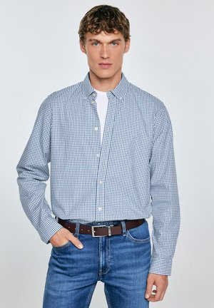 Camisa de cuadros azul y blanca, de manga larga, de algodón, usada sobre una camiseta blanca, combinada con jeans azules y un cinturón marrón.