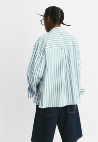 Persona con capelli intrecciati e occhiali, che indossa una camicia bianca larga con strisce verticali verdi e pantaloni corti di jeans scuri, rivolta verso di spalle su uno sfondo bianco.