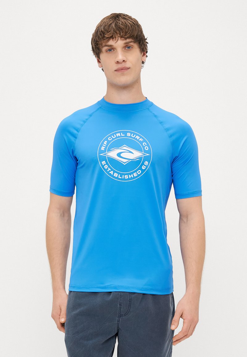 Zils īsām piedurknēm rash guards ar apļveida baltu logotipu uz priekšpuses, izgatavots no gludas, elastīgas materiāla, paredzēts valkāšanai ūdens aktivitātēs.