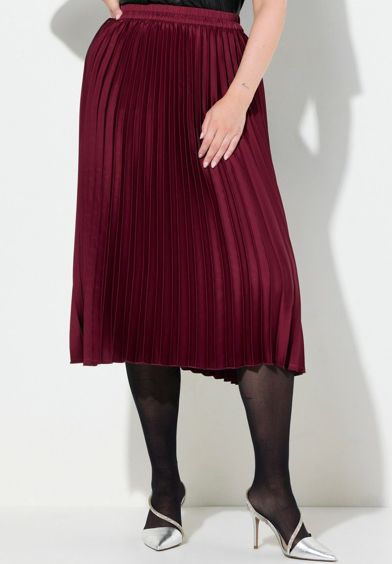 Burgundy plisserad midikjol med elastisk midja, kombinerad med genomskinliga svarta strumpbyxor och silverfärgade stilettklackar. Framhäver kroppens form och flöde.