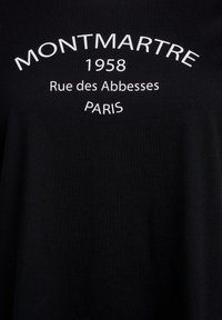 Crna pamučna majica s bijelim natpisom: "MONTMARTRE 1958 Rue des Abbesses PARIS." Jednostavan dizajn s okruglim izrezom.