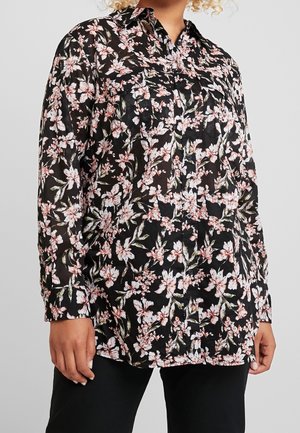 Zwarte blouse met lange mouwen en knopen, met roze en witte bloemenprint, gedragen over een zwarte broek, getoond van de middel tot de dijen.