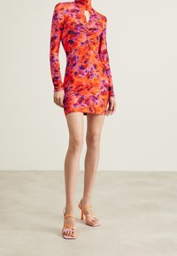 Robe ajustée en orange vif avec des motifs floraux en violet et rose. Présente des manches longues, un col haut et une découpe en forme de clé.