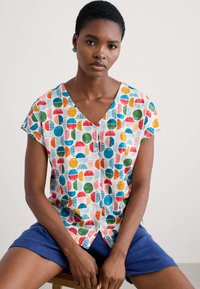 Short-sleeved, V-neck blouse with a colorful geometric pattern in red, blue, green, and yellow on a white background, made from lightweight fabric.