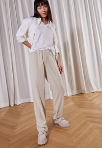 Beige lätta byxor med elastisk midja, i kombination med en vit V-halsad tröja och mjuka, ljusa skor. Trägolv i bakgrunden.