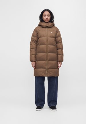 Kvinde står iført en brun dunfrakke til knæet med hætte, mørke vidde jeans og sorte slip-on sko mod en ensfarvet baggrund.