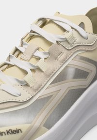 Κοντινή εικόνα του μπεζ και λευκού αθλητικού παπουτσιού με λευκά κορδόνια, σουέντ και διχτυωτές επιφάνειες, και μερικό λογότυπο Calvin Klein στη σόλα.