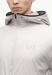 Άνδρας με ανοιχτό γκρι μπουφάν EA7 Emporio Armani με εμπρόσθιο φερμουάρ και προσαρτημένη κουκούλα, εμφανίζεται από τους ώμους έως το στόμα.
