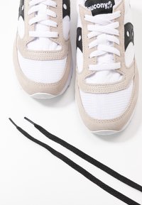 Paire de baskets Saucony blanches et beige avec logo noir et un jeu de lacets noirs placés en dessous sur une surface blanche.