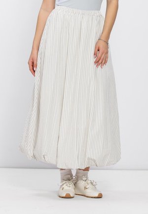 Vrouw draagt een witte en beige midi-rok met verticale strepen en plooien, met een elastische taille, gecombineerd met beige sokken en witte sneakers.