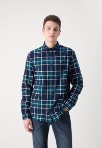 Camisa de cuadros azul marino con acentos verdes y blancos, con cuello abotonado y un logo de la marca sutil en el pecho.