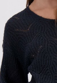 Close-up van een persoon die een zwart gebreid sweater draagt met een textuur en patronen, en een ronde halslijn.