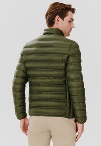 Zielona kurtka z ociepleniem, z poziomym pikowaniem, stójką i elastycznymi mankietami. Materiał wydaje się lekki i gładki w dotyku.