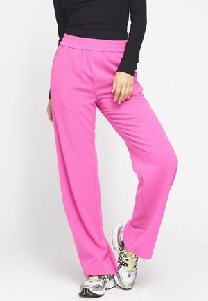 Person trägt leuchtend rosa weite Hosen, schwarzes Langarmshirt und weiße Laufschuhe mit neongelben Schnürsenkeln, steht auf weißem Boden.