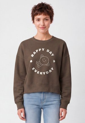 Jeune femme portant un sweat-shirt marron avec un visage souriant et le texte "Happy Day Everyday,", associé à un jean bleu clair, debout sur fond blanc.