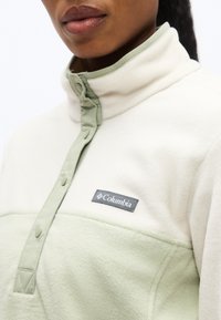 Κοντινό πλάνο ατόμου που φοράει δίχρωμο φλις πουλόβερ σε ανοιχτό πράσινο με το λογότυπο Columbia στο στήθος και γιακά με κουμπιά τύπου snap.