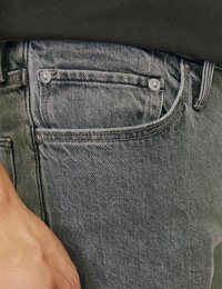 Šedé džíny z denimu s texturovaným povrchem, obsahující malou přední kapsu a dva kovové knoflíky na pasu. Ruka spočívá na látce.