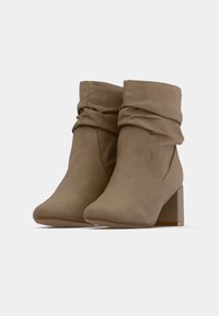 Béžové členkové topánky so zmäkčeným dizajnom, vyrobené z mäkkého materiálu. Majú blokový podpätok a bezšvové bočné detaily pre uhladený vzhľad.