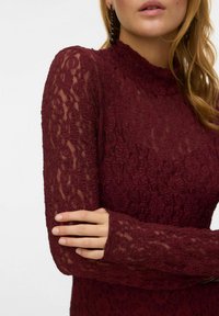 Viininpunainen pitsitoppi pitkällä, läpikuultavalla hihalla, korkealla kaula-aukolla, teksturoidulla kukkakuviolla ja istuvilla hihansuihilla. Sileä pinta ja yksityiskohtainen muotoilu.