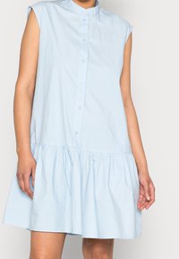 Vaaleansininen nappikauluspaita, jossa on röyhelöhelma, lyhyet hihat ja korkea kaulus. Valmistettu sileästä, kevyestä kankaasta.