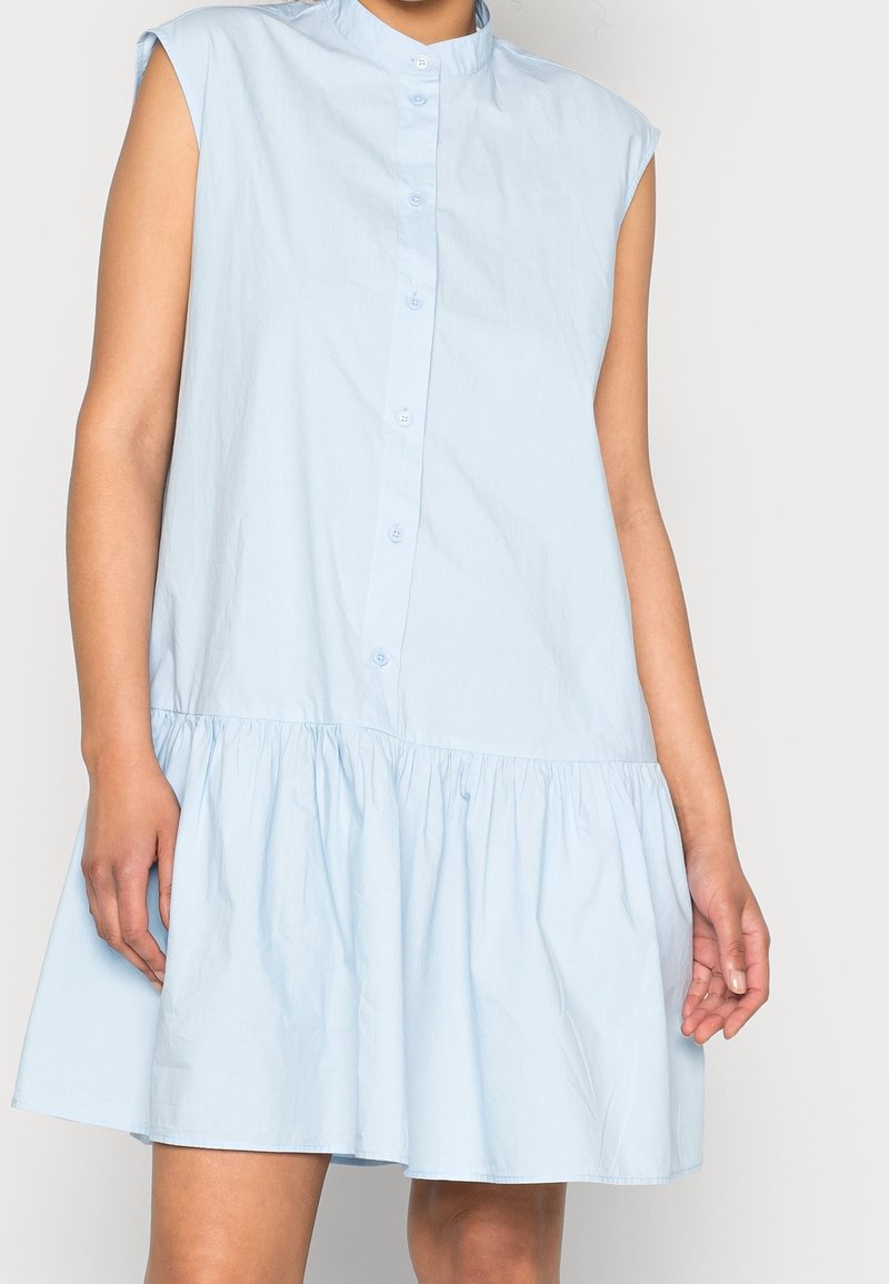 Vaaleansininen nappikauluspaita, jossa on röyhelöhelma, lyhyet hihat ja korkea kaulus. Valmistettu sileästä, kevyestä kankaasta.